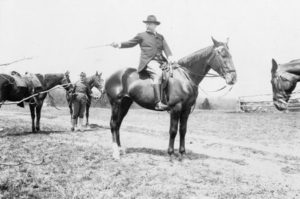 Black and white photo of Theodore Roosevelt on a horse, selecting the next member of the outing party for the next jump over the hurdles, 1907.