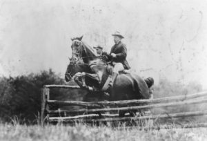 Black and white photo of Theodore Roosevelt and Capt Fitzhugh on horses, jumping a fence, 1907.